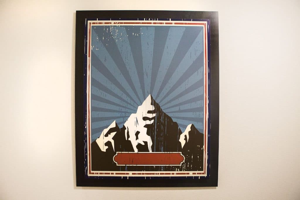 A framed poster with a stylized illustration of a snow-capped mountain against a sky with radial lines. The bottom features a blank, horizontal red label.