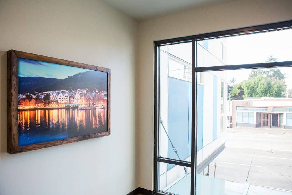 A framed picture of a lit-up town by the water hangs on a white wall next to large windows overlooking a street.