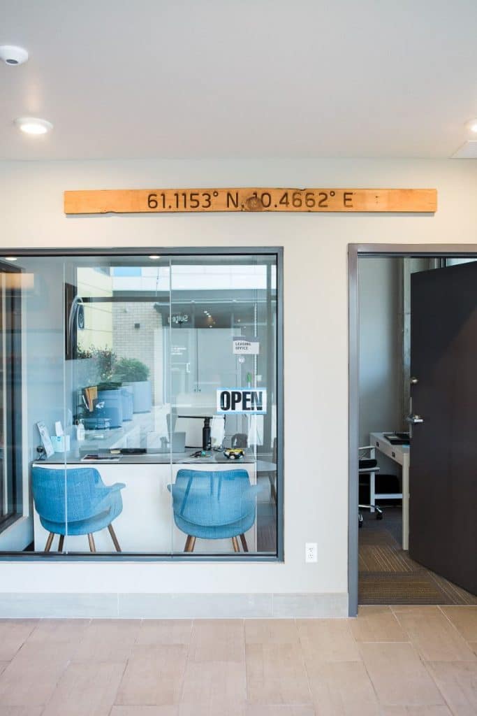 Modern office entrance with a glass window showing a room with blue chairs and a desk. A wooden sign displaying coordinates hangs above. An "OPEN" sign is visible in the window.