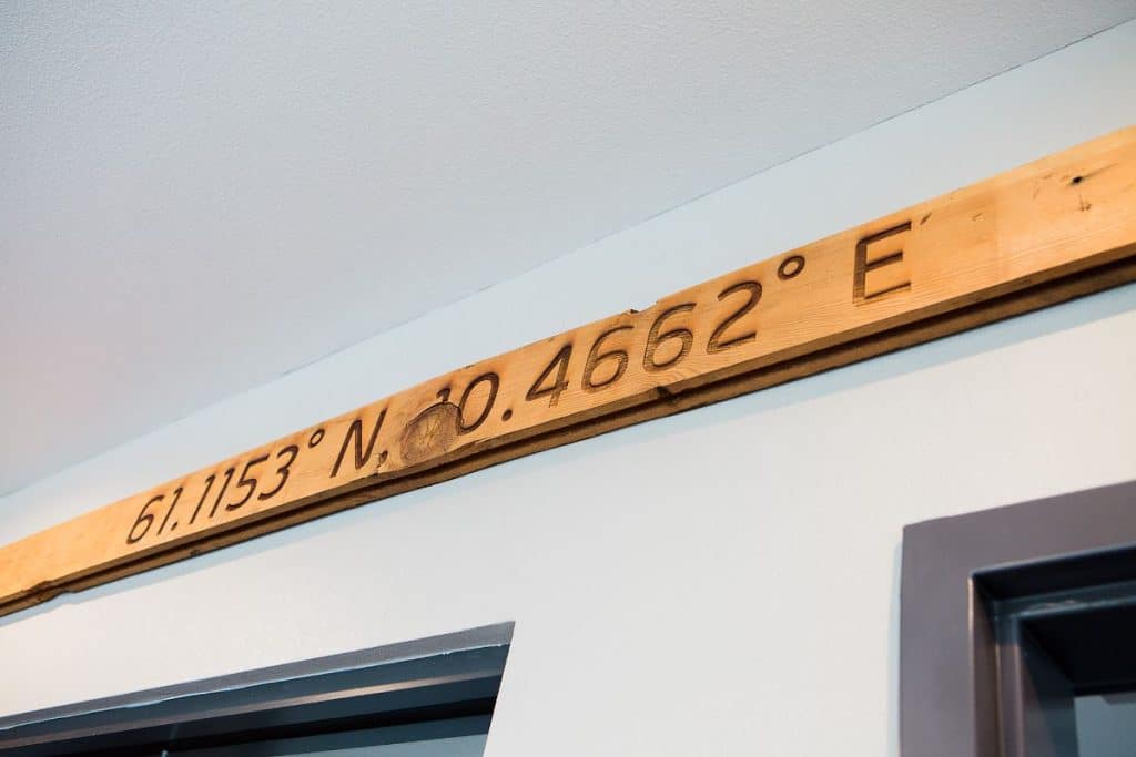 Wooden beam displaying the coordinates 61.1153° N, 10.4662° E mounted on a light blue wall.