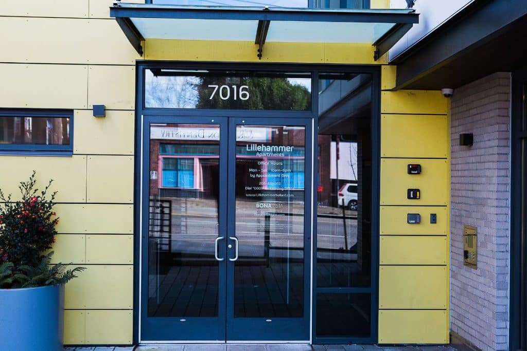 Glass door entrance with the number 7016 above. Yellow paneling and a white brick wall are visible. Reflections of buildings and a parked car can be seen on the door.