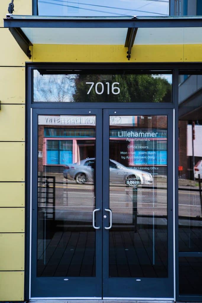 Glass door of an apartment building with the address number 7016 displayed above. Reflections of a car and buildings can be seen in the glass.