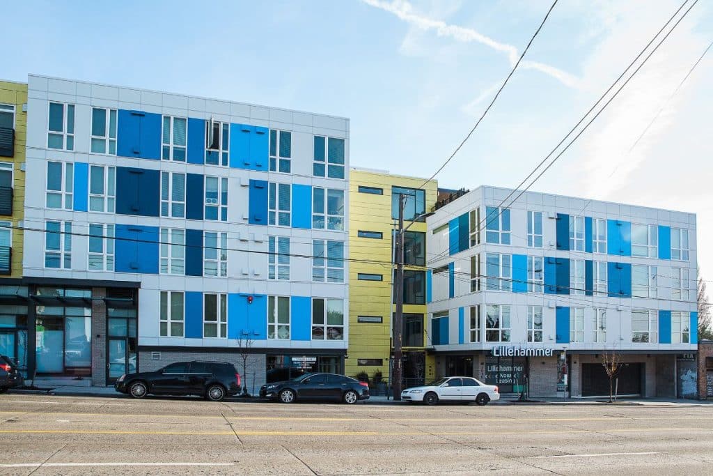 Modern apartment complex with colorful panels, featuring multiple stories and large windows, situated along a street with parked cars.