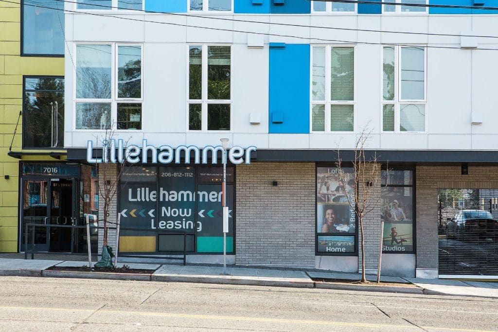 Street view of a building with "Lillehammer Now Leasing" sign and glass doors showing interior posters advertising lofts, studios, and home amenities.