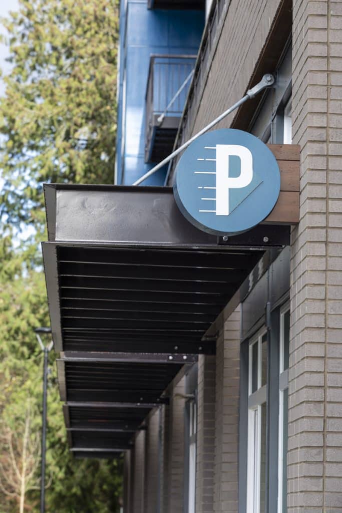 Building facade with a round blue parking sign displaying a white 'P' under a metal awning. Trees are visible in the background.