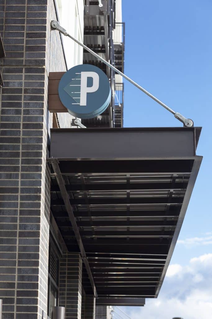 A circular blue parking sign with a white "P" is mounted above a metal awning on a brick building, set against a clear blue sky.