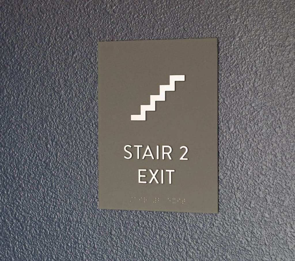 Sign reading "Stair 2 Exit" with an icon of stairs and Braille text below, mounted on a textured gray wall.