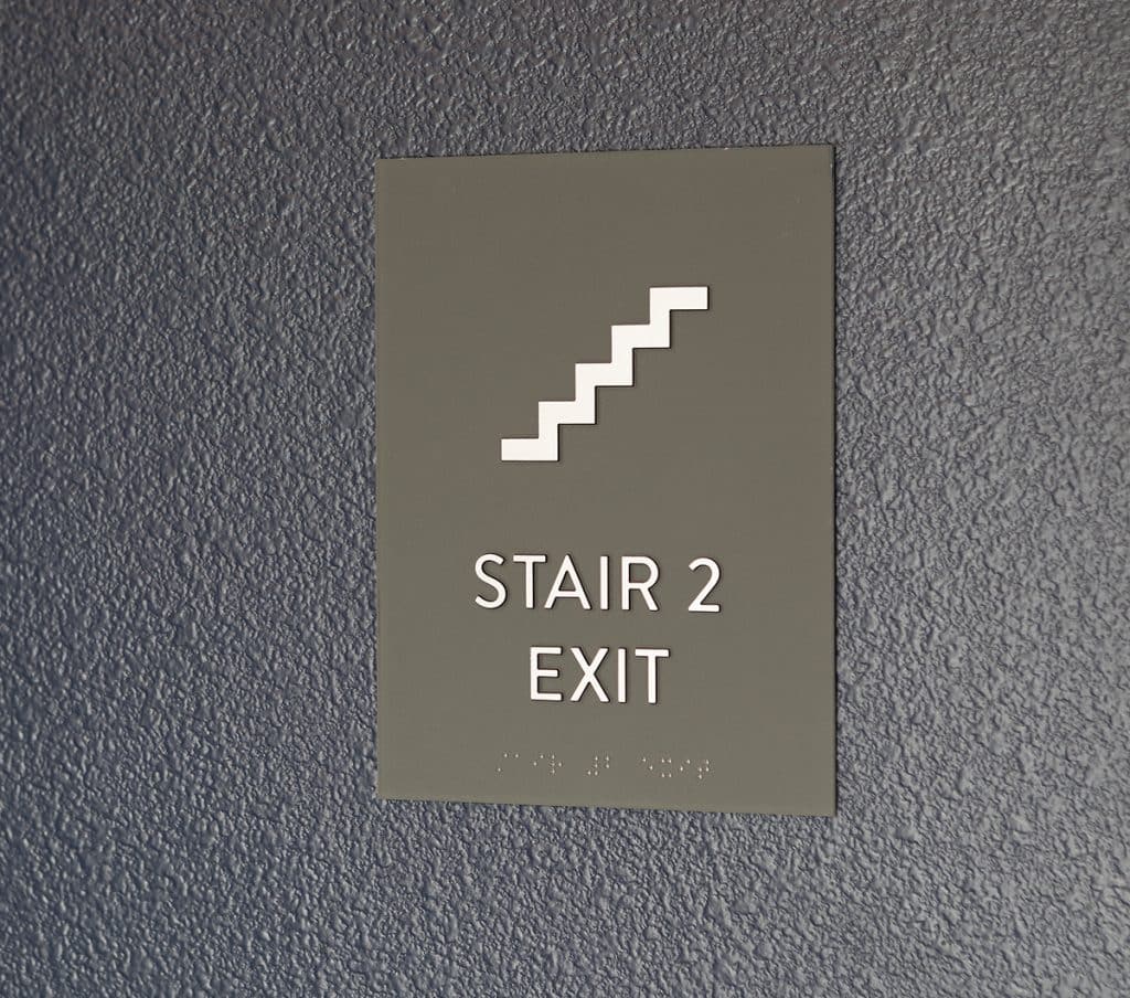 Sign on a textured wall reads "Stair 2 Exit" with a stair icon above. Braille is present below the text.