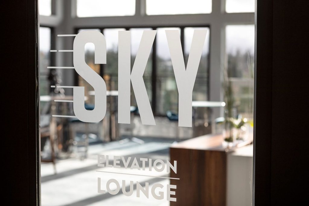 Glass door with "Sky Elevation Lounge" written on it, overlooking a bright room with large windows and modern decor.