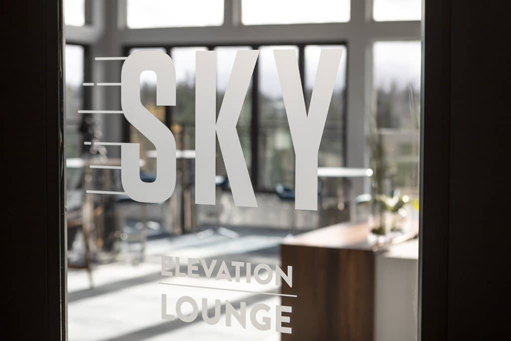 Glass door with "SKY Elevation Lounge" text, overlooking a bright room with large windows and modern decor.