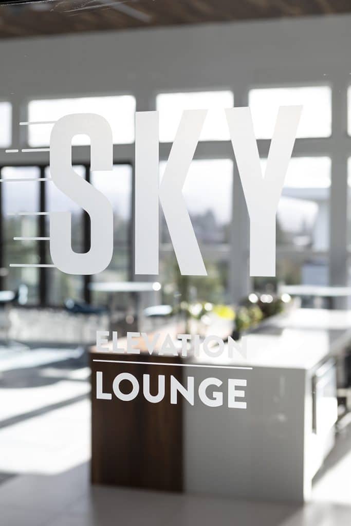 Text on glass reads "Sky Elevation Lounge" with a view of a modern interior and large windows in the background.