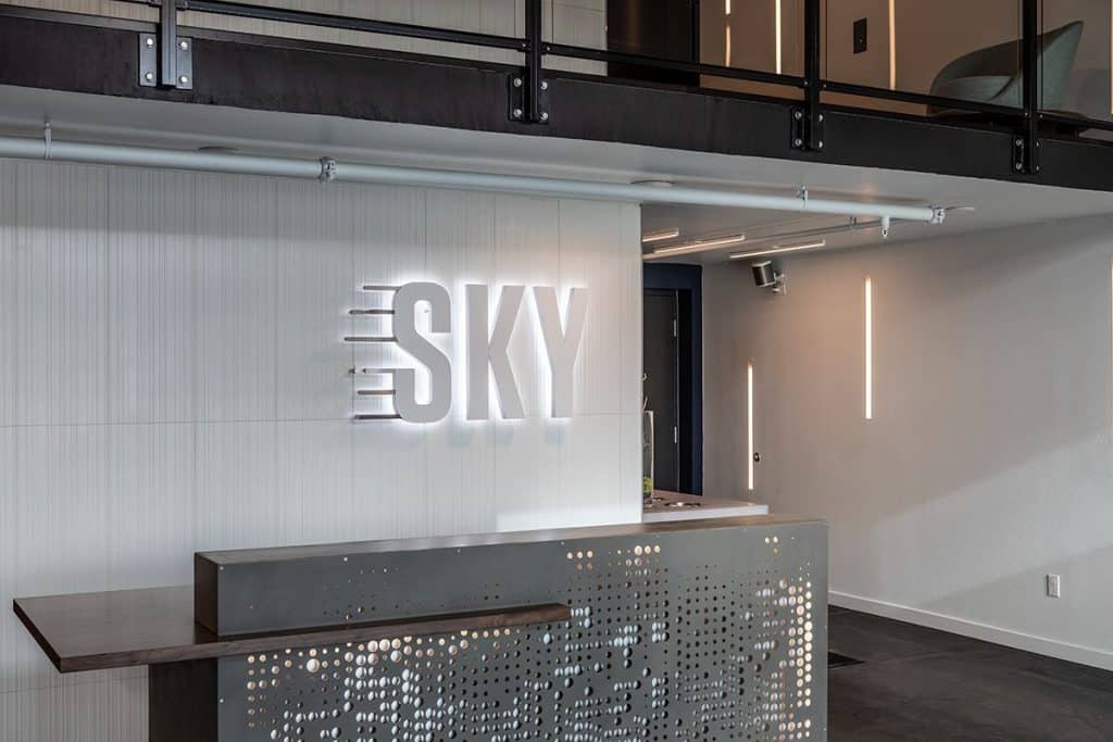 Reception desk with perforated metal front and illuminated "SKY" sign on the wall behind. Modern design with minimal decor.