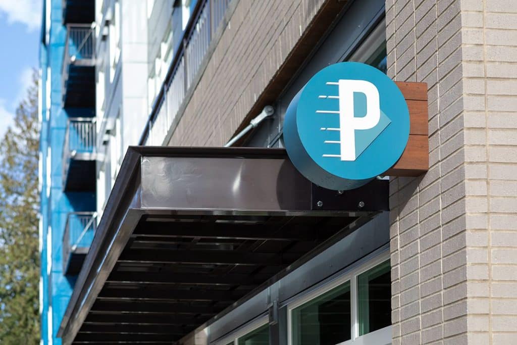 A circular sign with a white "P" and lines on a blue background is mounted on a brick building near a metal awning.