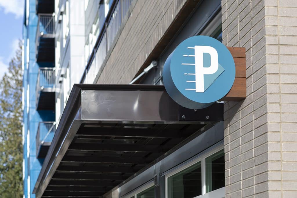 Blue "P" parking sign on a building's exterior with windows and a tree in the background.