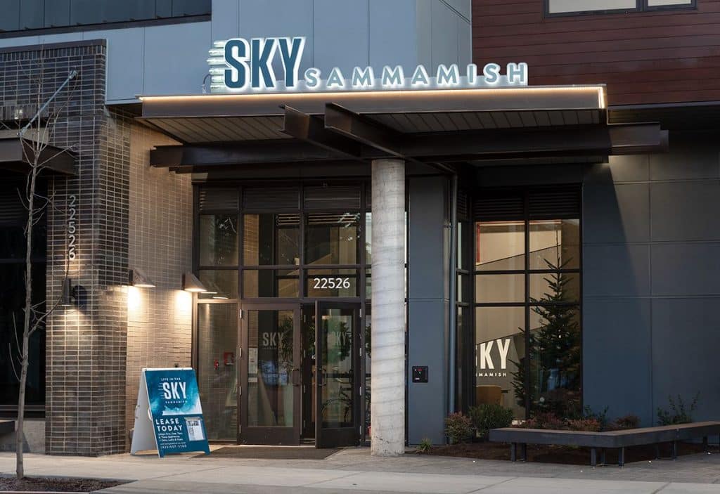 Modern building entrance with glass doors, "Sky Sammamish" sign above, and promotional signboard on the left.