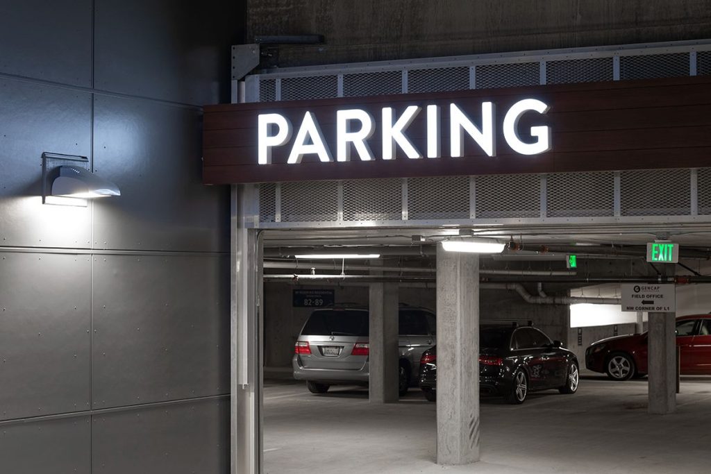 Underground parking garage entrance with illuminated "PARKING" sign. Several cars parked inside under fluorescent lighting. Exit sign visible on the right.