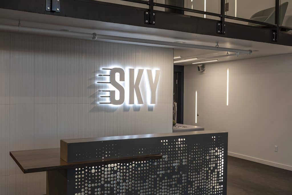 Reception area with a modern design. The wall has a glowing "SKY" sign, and the desk features a pattern of small circular cutouts. Neutral tones and minimalist lighting are used.