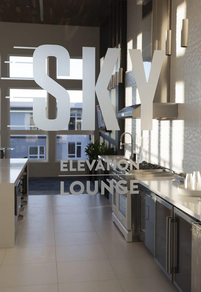 View of a modern kitchen area with large windows, labeled 'SKY ELEVATION LOUNGE' on the glass, showing countertops, appliances, and pendant lights.