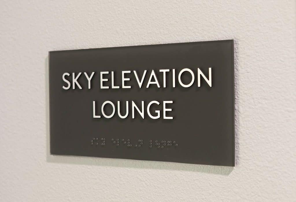 A sign reading "Sky Elevation Lounge" with braille text below, mounted on a wall.