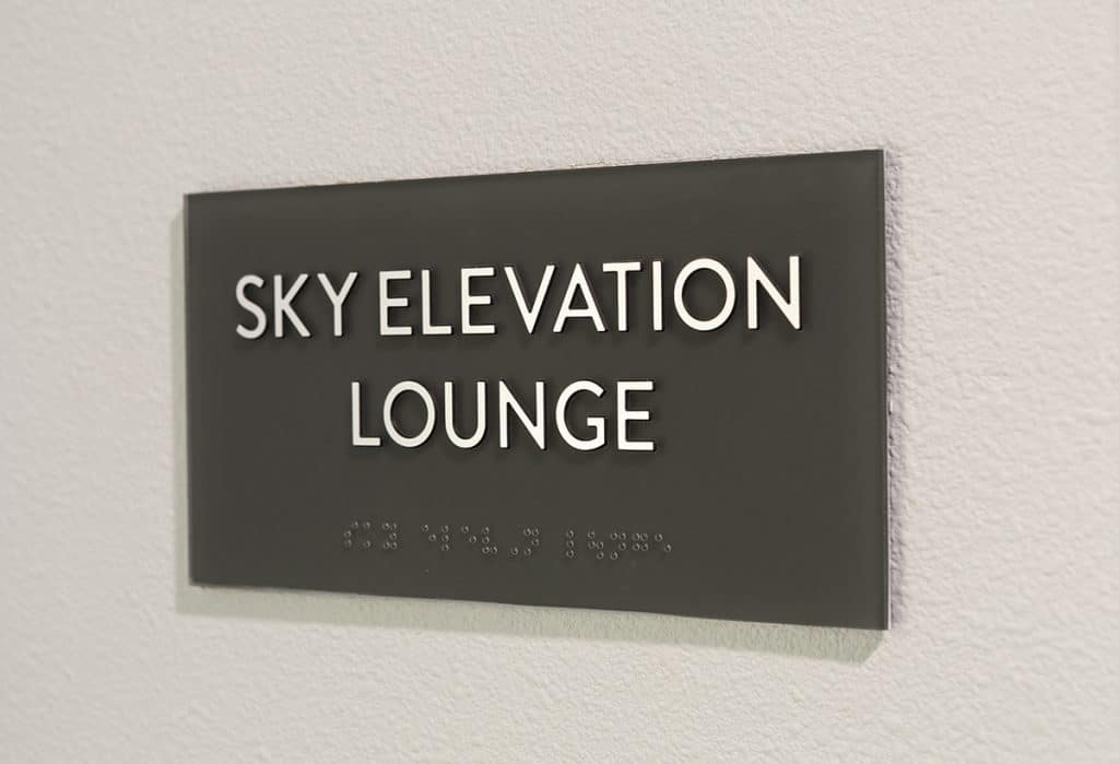 Sign on a wall reads "Sky Elevation Lounge" with braille beneath the text.