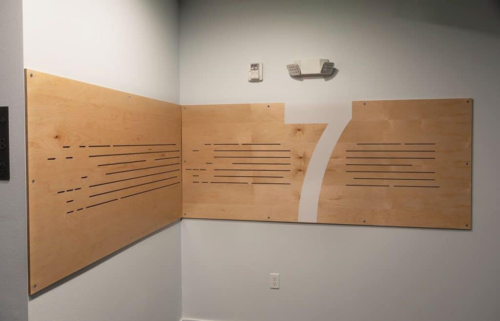 Wood panels with horizontal slits form a corner installation on a wall. A light fixture and outlet are above and below the panels. One panel has a white number "7" design.