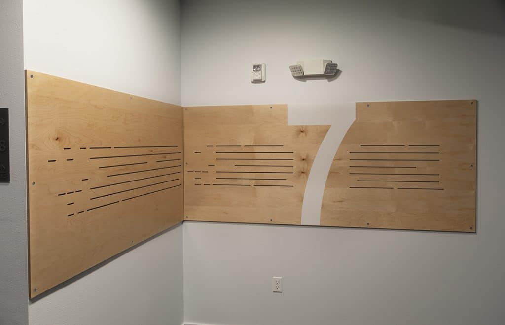 Wooden wall panels with horizontal slits form the number 7. White wall, overhead lights, and electrical outlet are visible.