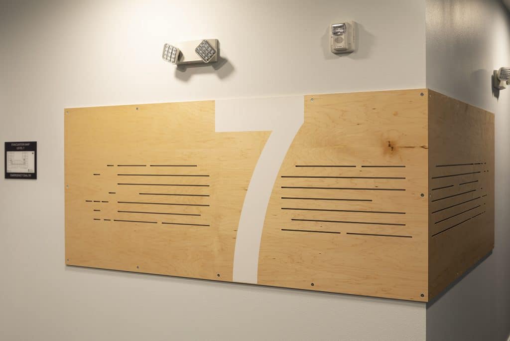 Wooden wall sign with the number 7 and horizontal line cutouts, mounted on a corner under two wall lights.