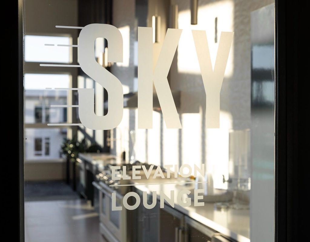 Glass door with "Sky Elevation Lounge" text, revealing a modern interior with large windows and sleek countertops.