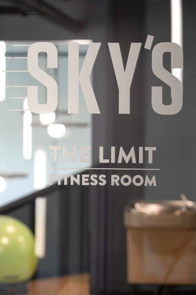 Sign on a glass door reads, "SKY'S THE LIMIT FITNESS ROOM," with blurred interior, exercise ball, and water fountain visible in the background.