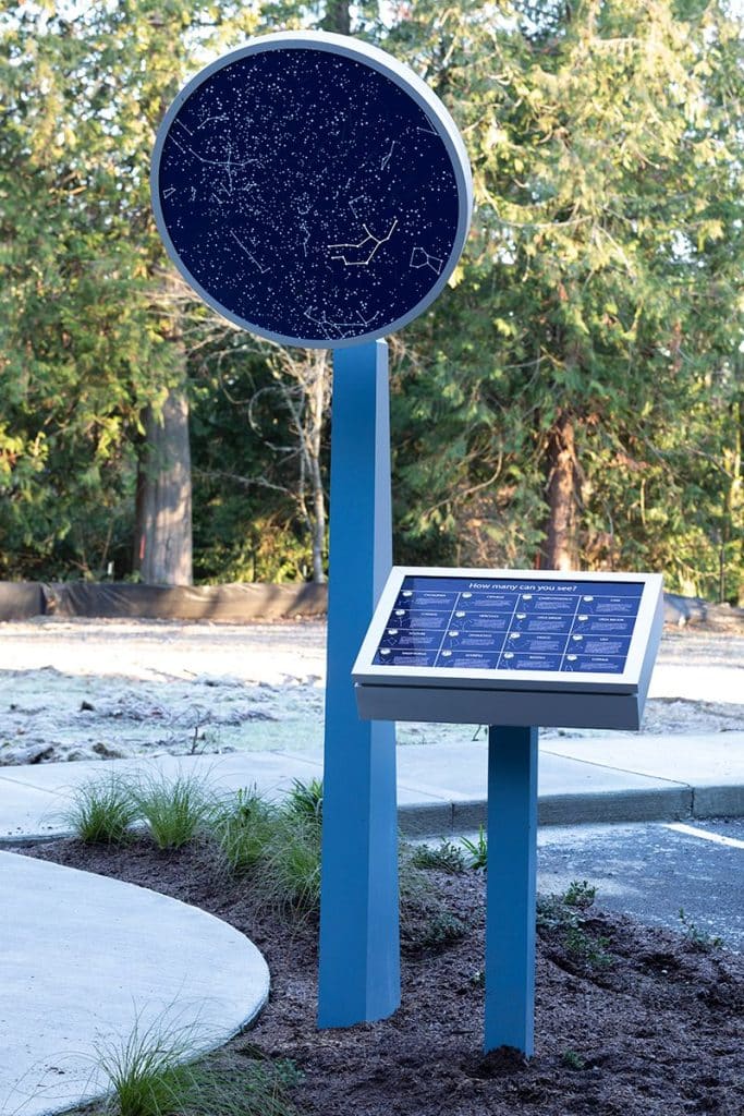 A blue star map display in a park showing constellations, accompanied by an information board, surrounded by trees and pathways.