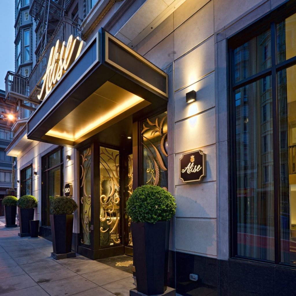 Entrance of a restaurant with a modern design, featuring a sign with illuminated lettering, large windows, decorative glass doors, and potted topiary plants.