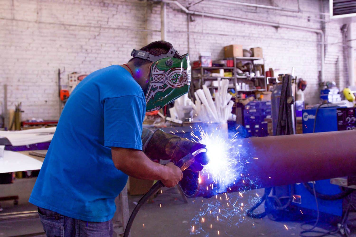 Person wearing a welding helmet and blue shirt, welding a large metal pipe in an industrial workshop setting. Shelves and equipment in the background.