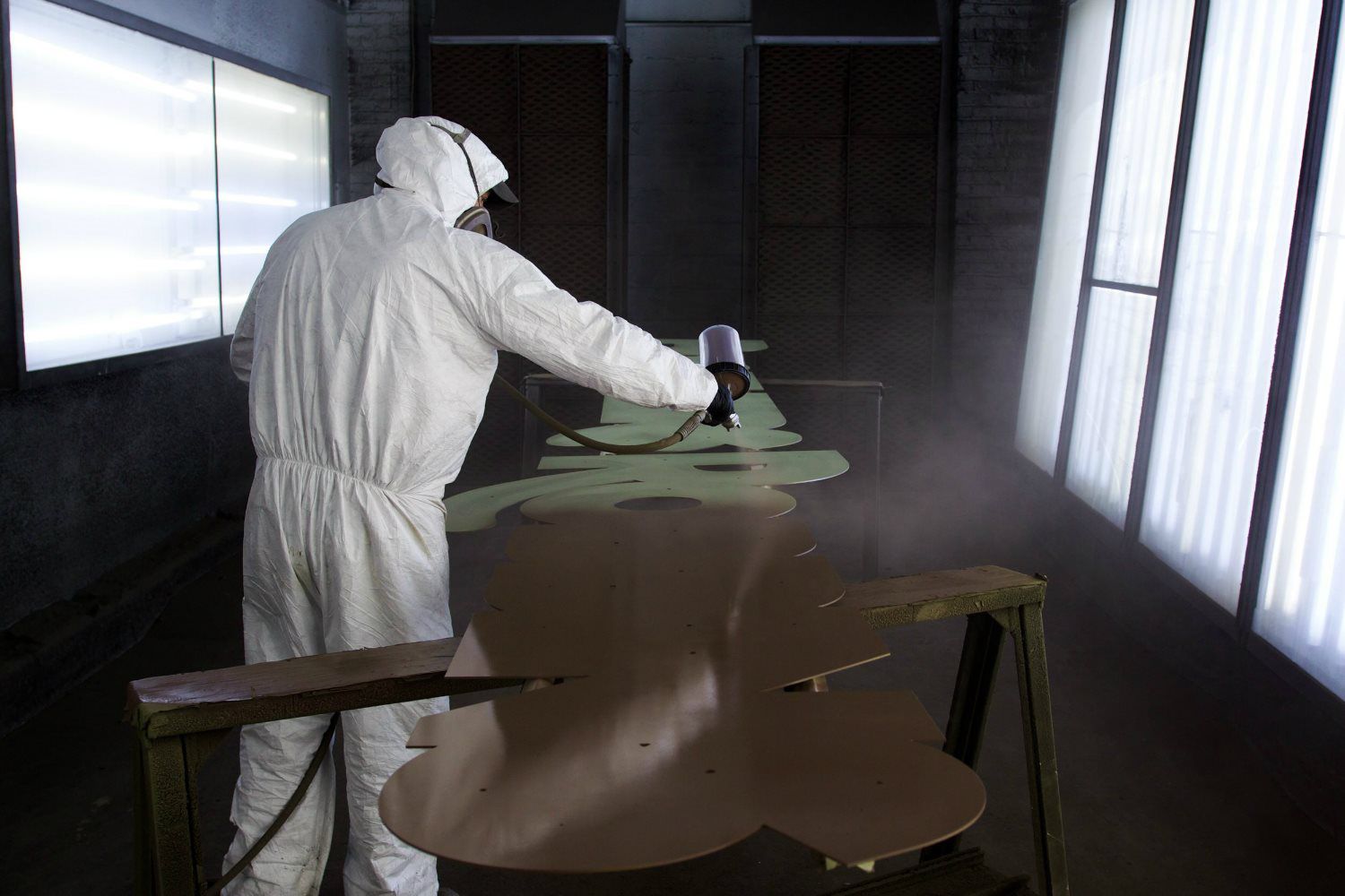 A person in protective gear spray paints large metal parts in a dimly lit room.