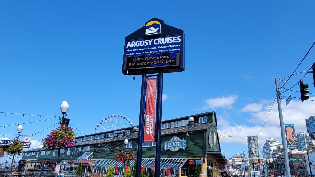 Sign for Argosy Cruises with a ferris wheel in the background, adjacent to a green building with "Elliott's" signage. Bright blue sky and potted flowers visible.