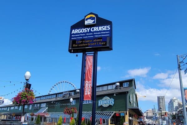 Sign for Argosy Cruises with a ferris wheel in the background, adjacent to a green building with "Elliott's" signage. Bright blue sky and potted flowers visible.