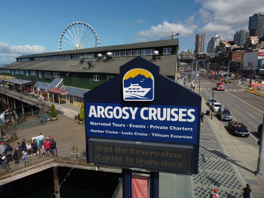 Argosy Cruises sign with information about narrated tours and charters, set against a backdrop of a waterfront building, ferris wheel, and city street.