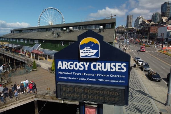 Argosy Cruises sign with information about narrated tours and charters, set against a backdrop of a waterfront building, ferris wheel, and city street.