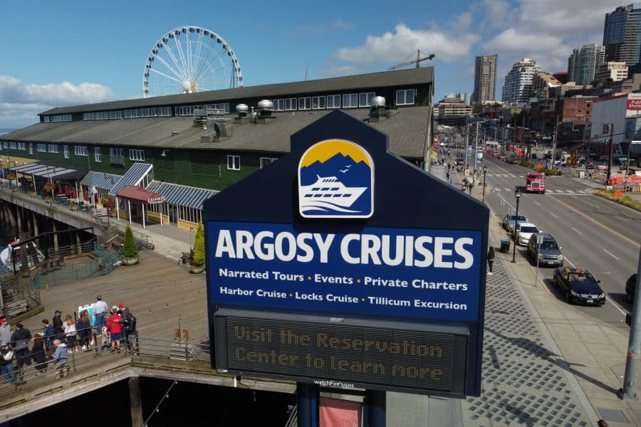 Argosy Cruises sign with information about narrated tours and charters, set against a backdrop of a waterfront building, ferris wheel, and city street.