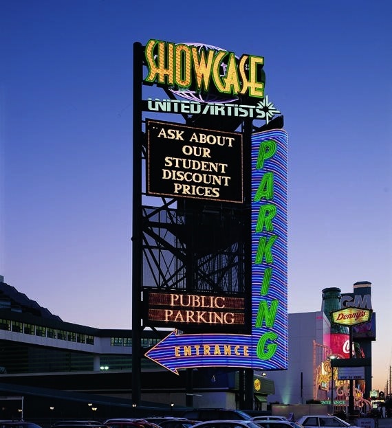 Neon sign for Showcase United Artists advertising student discount prices, public parking, and entrance with arrow, set against a twilight sky.