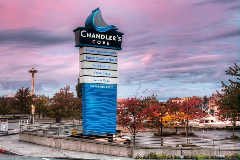 Chandler's Cove sign with business listings against a pink sky backdrop. Nearby foliage is turning autumnal, and the Space Needle is visible in the background.