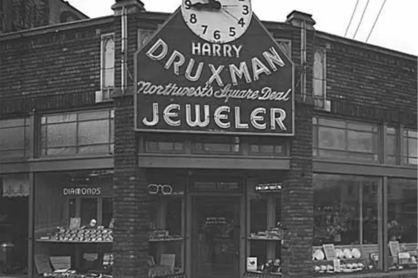 Black and white photo of a storefront with a large clock above the sign "Harry Druxman Northwest's Square Deal Jeweler." Display windows show various jewelry items.