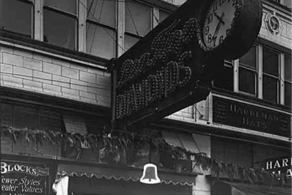 Black and white photo of a building facade with a vintage double-sided clock and sign advertising diamonds. Roofline shows "Block's Fewer Styles Greater Values" and "Hardeman Hats.