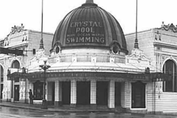 A historic building with a large dome labeled "Crystal Pool," featuring signs for swimming and warm ocean water, set against a cloudy sky.