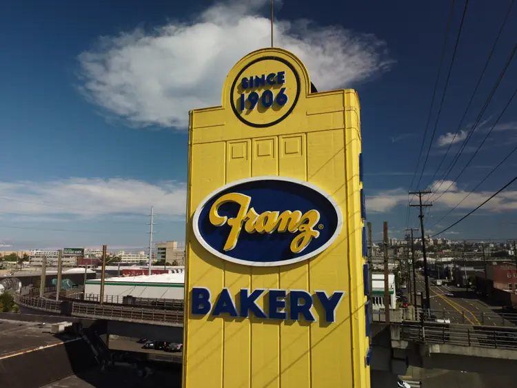 Tall yellow sign for Franz Bakery, established in 1906, with industrial background and road nearby.