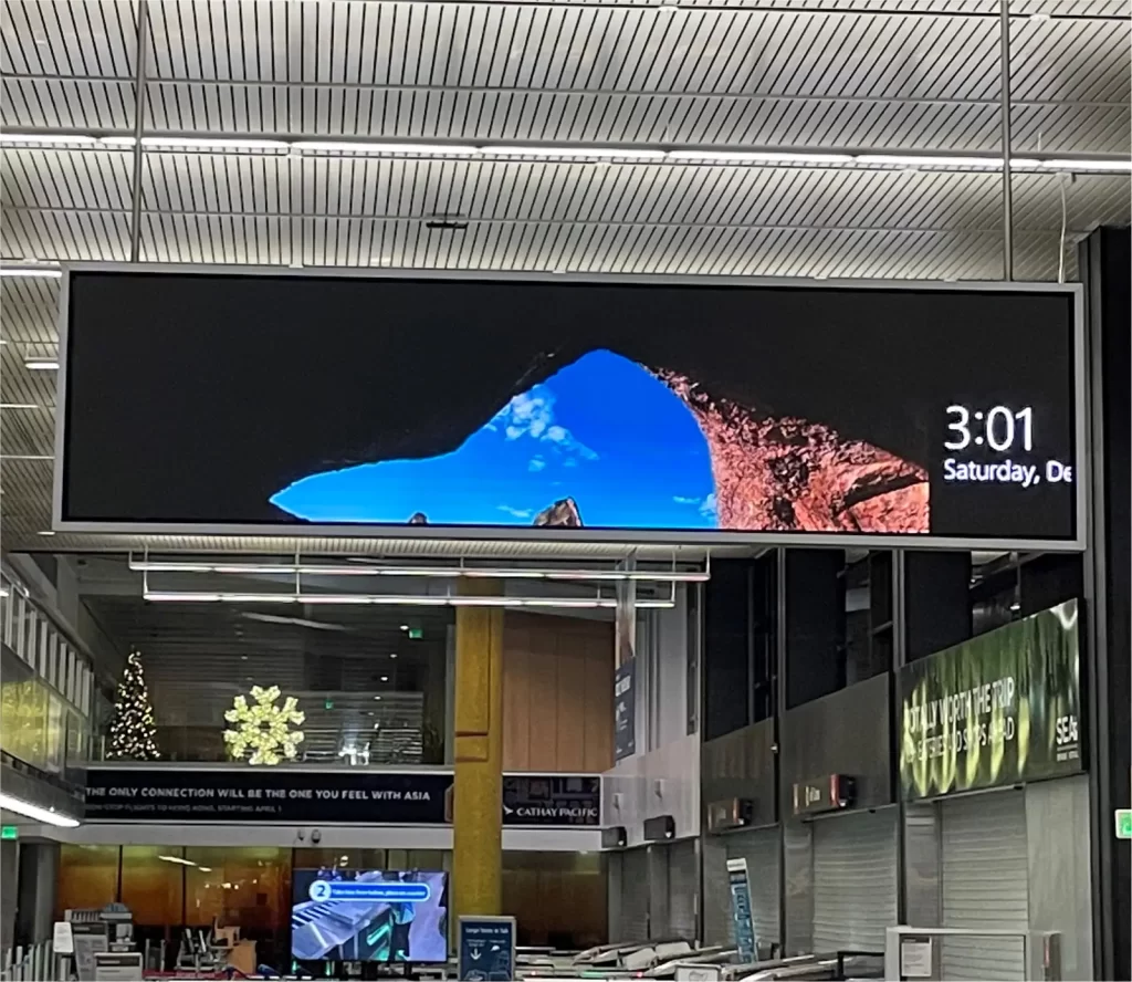 Digital clock display in an airport shows the time as 3:01 PM on Saturday, with a scenic landscape image above. Terminal features holiday decorations and airline signs.