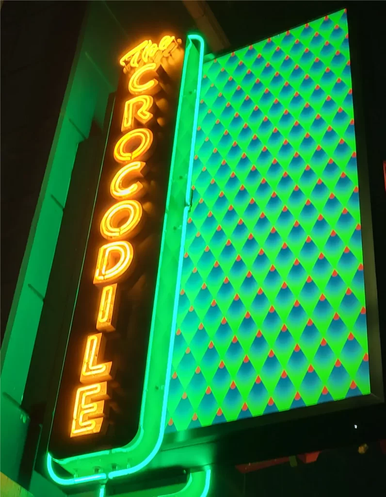 Neon sign of "The Crocodile," featuring bold glowing text and a vibrant geometric pattern in green and blue.