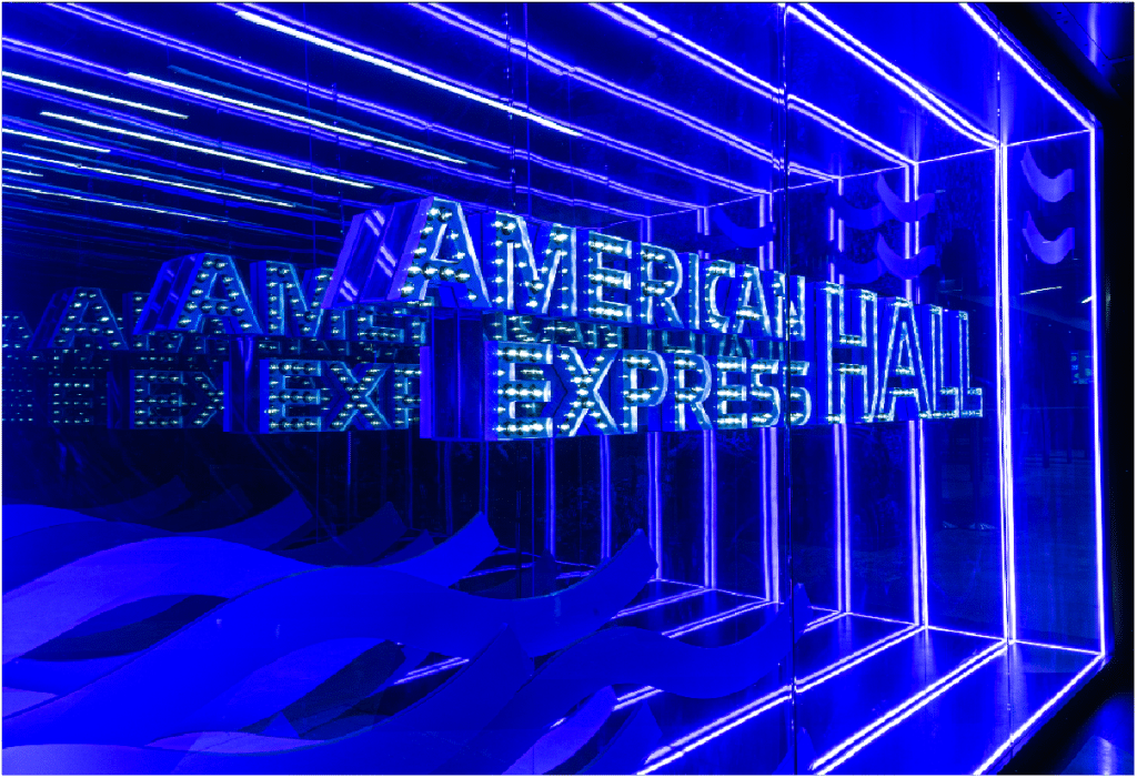 Blue neon sign reading "AMERICAN EXPRESS HALL" with reflections in a mirrored space.