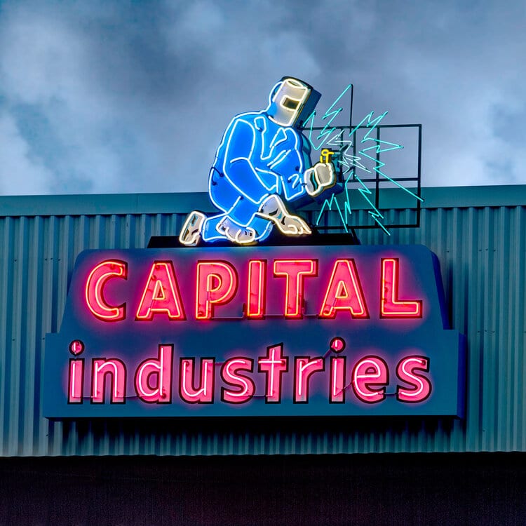 Neon sign for "Capital Industries" featuring a blue welder with a torch and sparks.