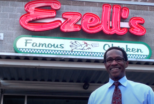 A person in a shirt and tie stands smiling in front of the Ezell's Famous Chicken restaurant, with its neon sign visible above.