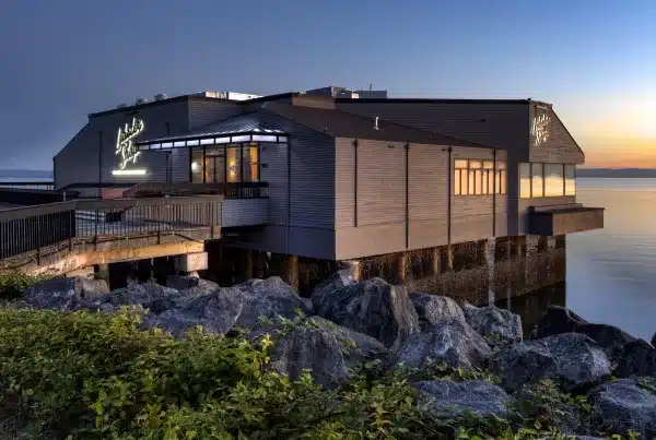 A gray building with a neon sign extends over a rocky shoreline at sunset, with calm water in the background.